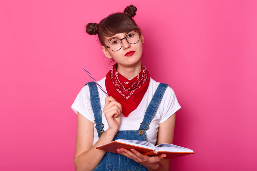 A young woman with glasses and double buns, wearing overalls and a red bandana, holds an open book and a pencil against a pink background, exuding a quirky, bookish persona.