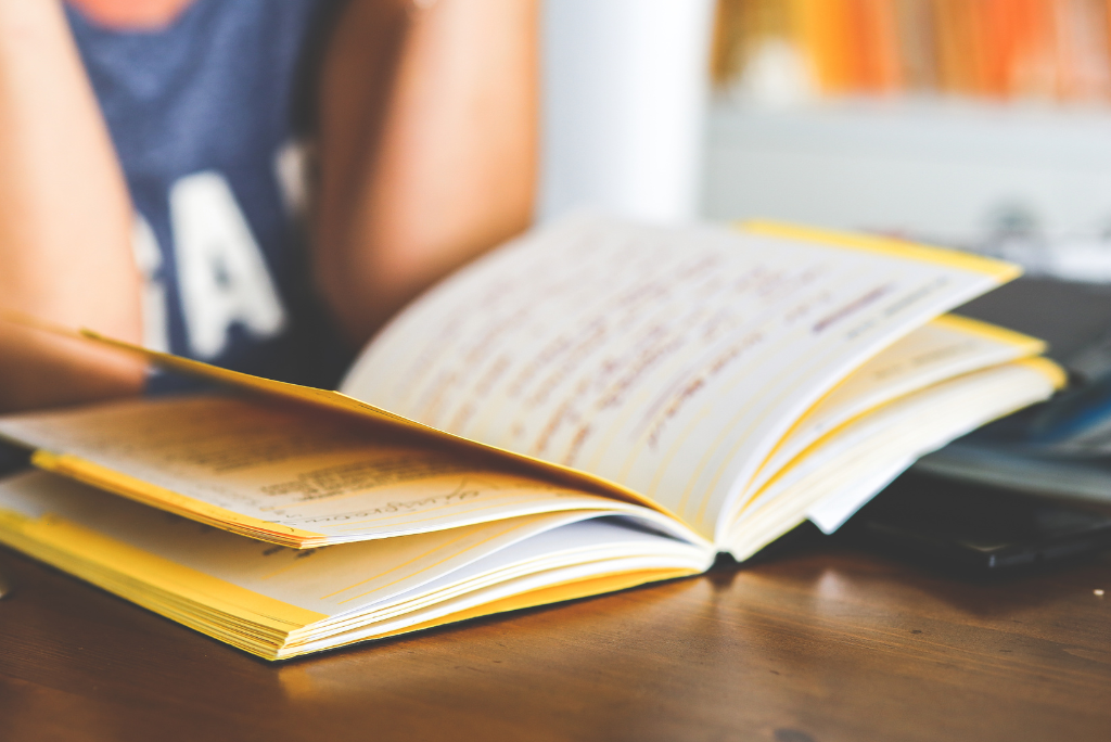 Close-up of an open notebook filled with handwriting on a wooden table, with a person sitting nearby, suggesting summer journaling or reading notes.