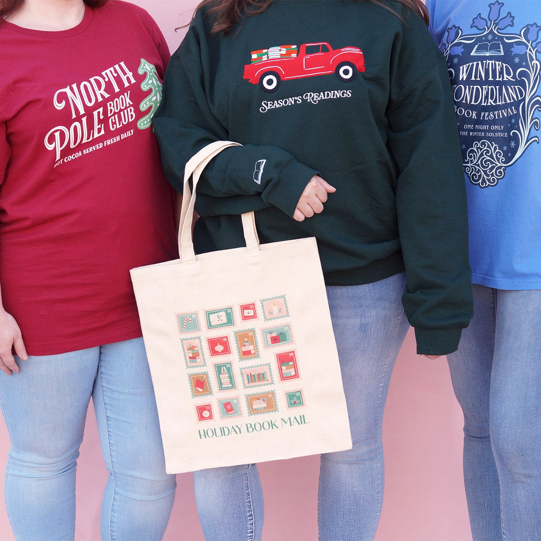 three people wearing bookish winter themed apparel and holding a holiday book mail tote bag.