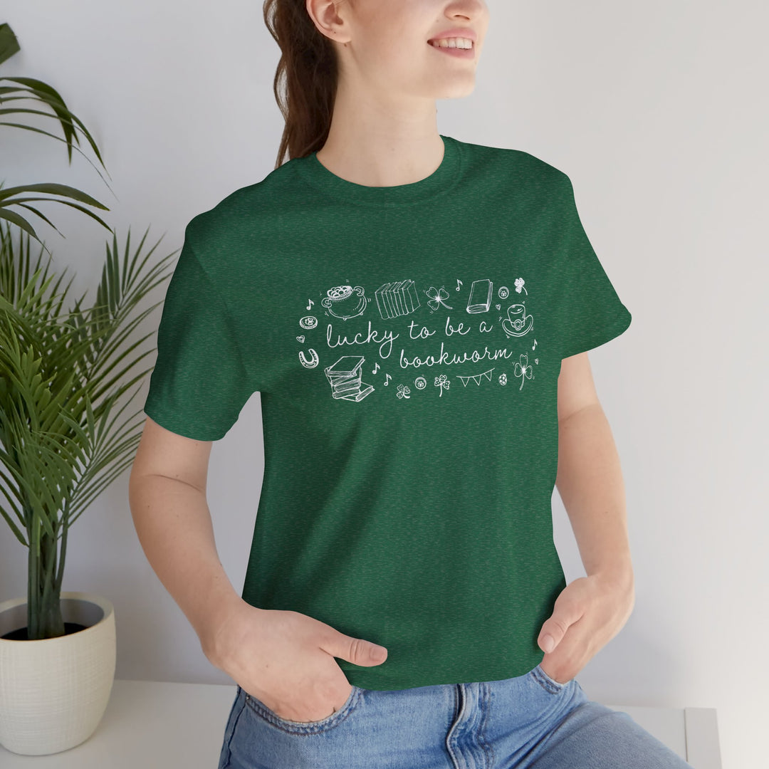 Person wearing a green t-shirt with white font reading "lucky to be a bookworm" and line illustrations of St. Patrick's Day items and books standing indoors next to a plant.