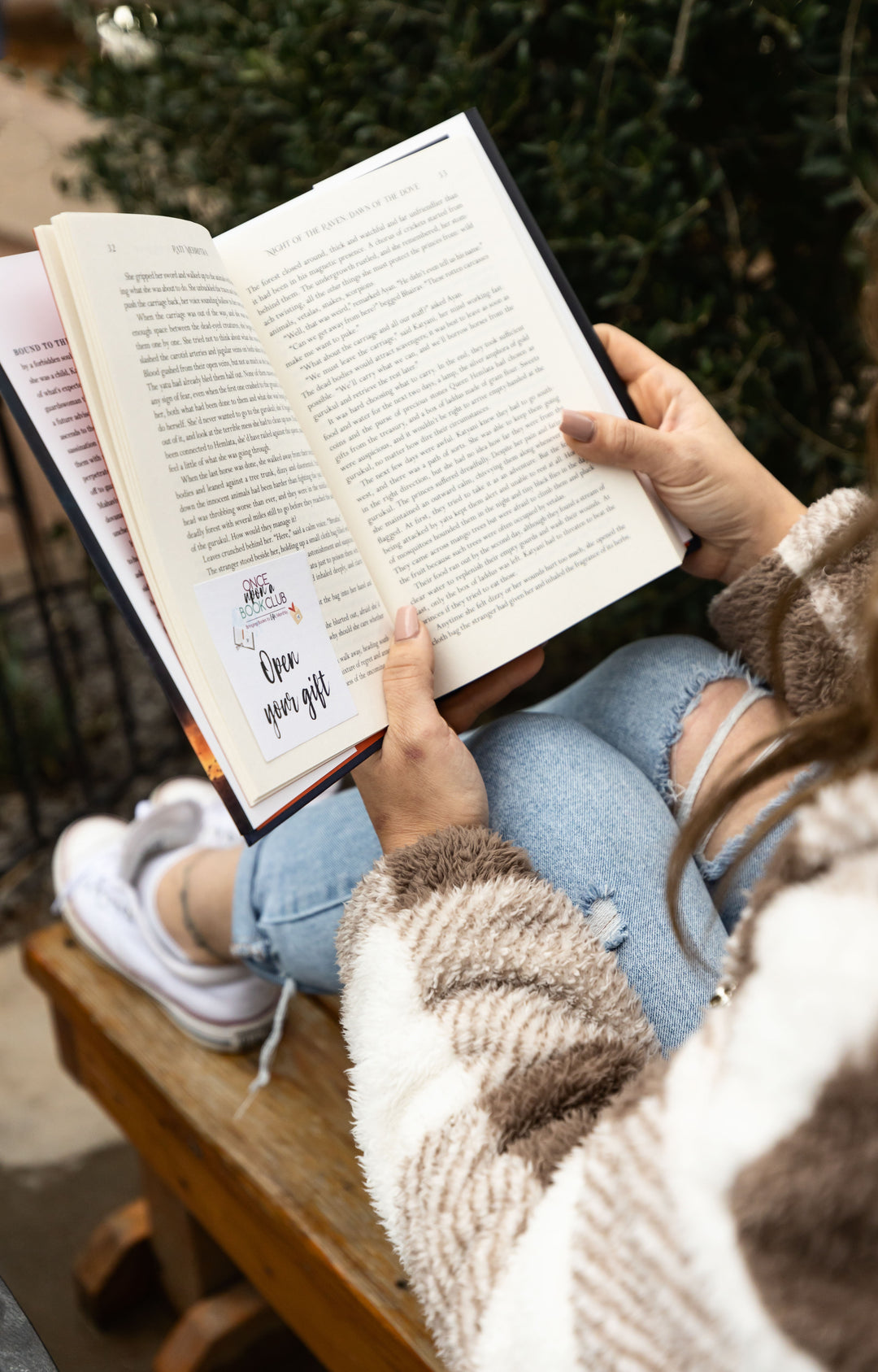 Person reading a book outdoors with greenery in the background. An "open your gift" sticky note can be seen inside the book.