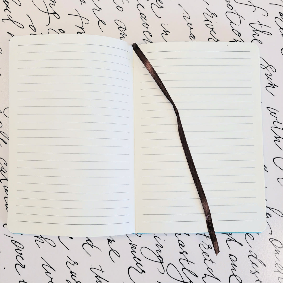 A lined notebook sits open on a white background decorated with script. The notebook has a fabric bookmark draped over the right page.