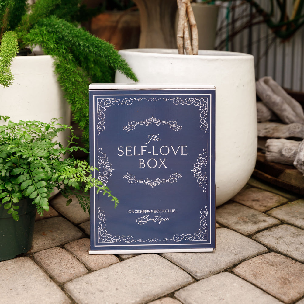 A navy blue Self-Love Box sits outdoors surrounded by greenery.