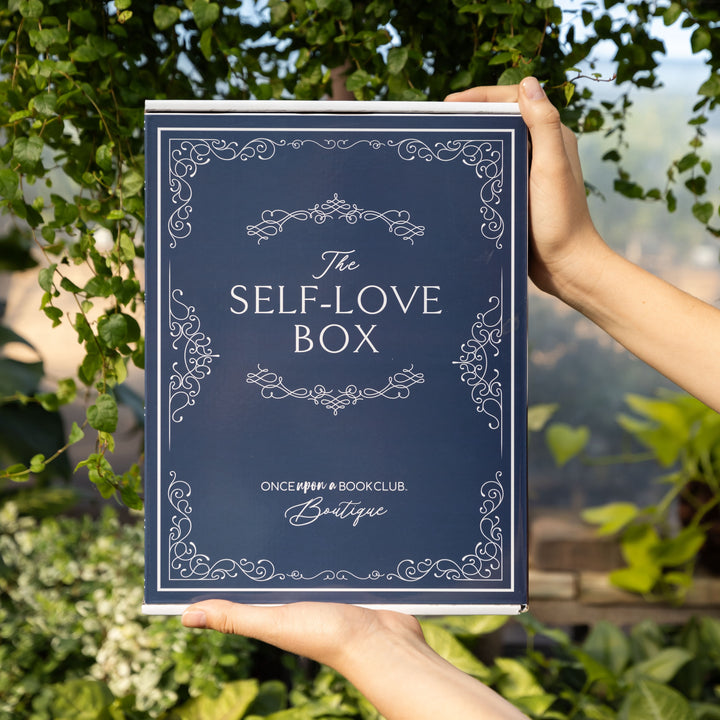 A navy blue box is held by a pair of hands in front of lush greenery.