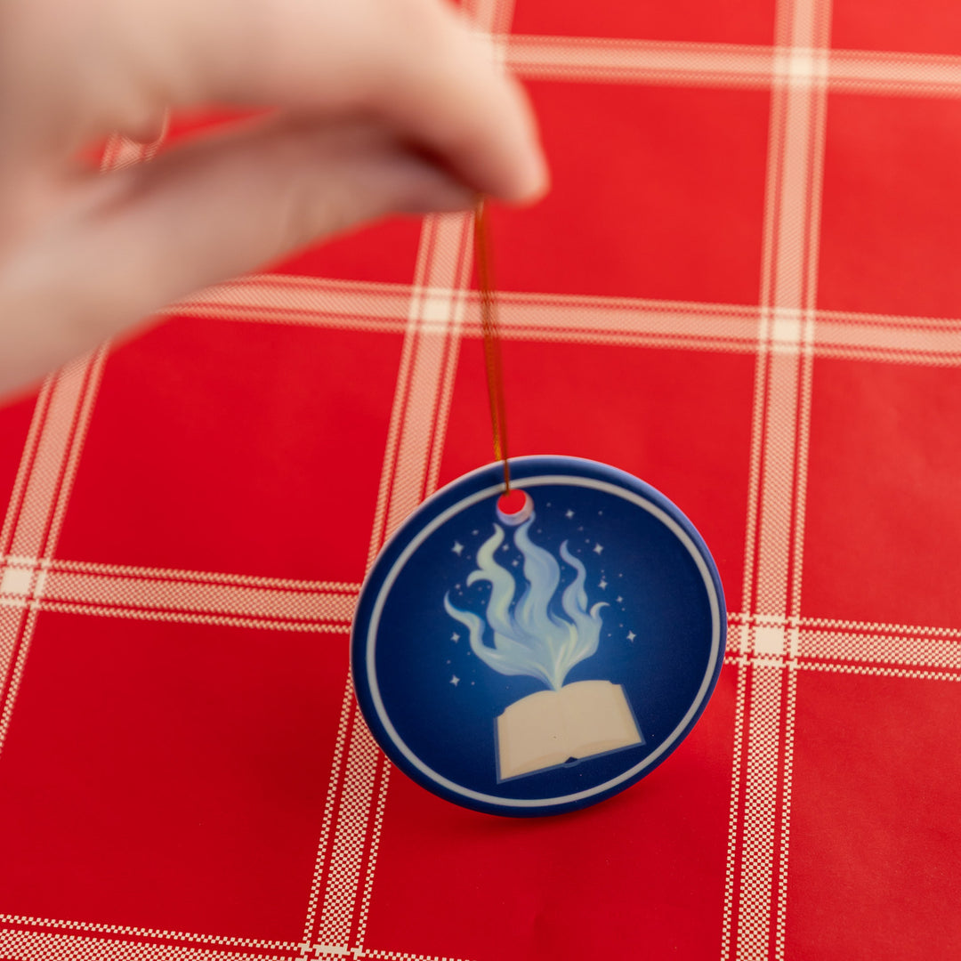 A hand holds a circular ornament that shows an open book with whisps of magic coming out from the center of the pages.