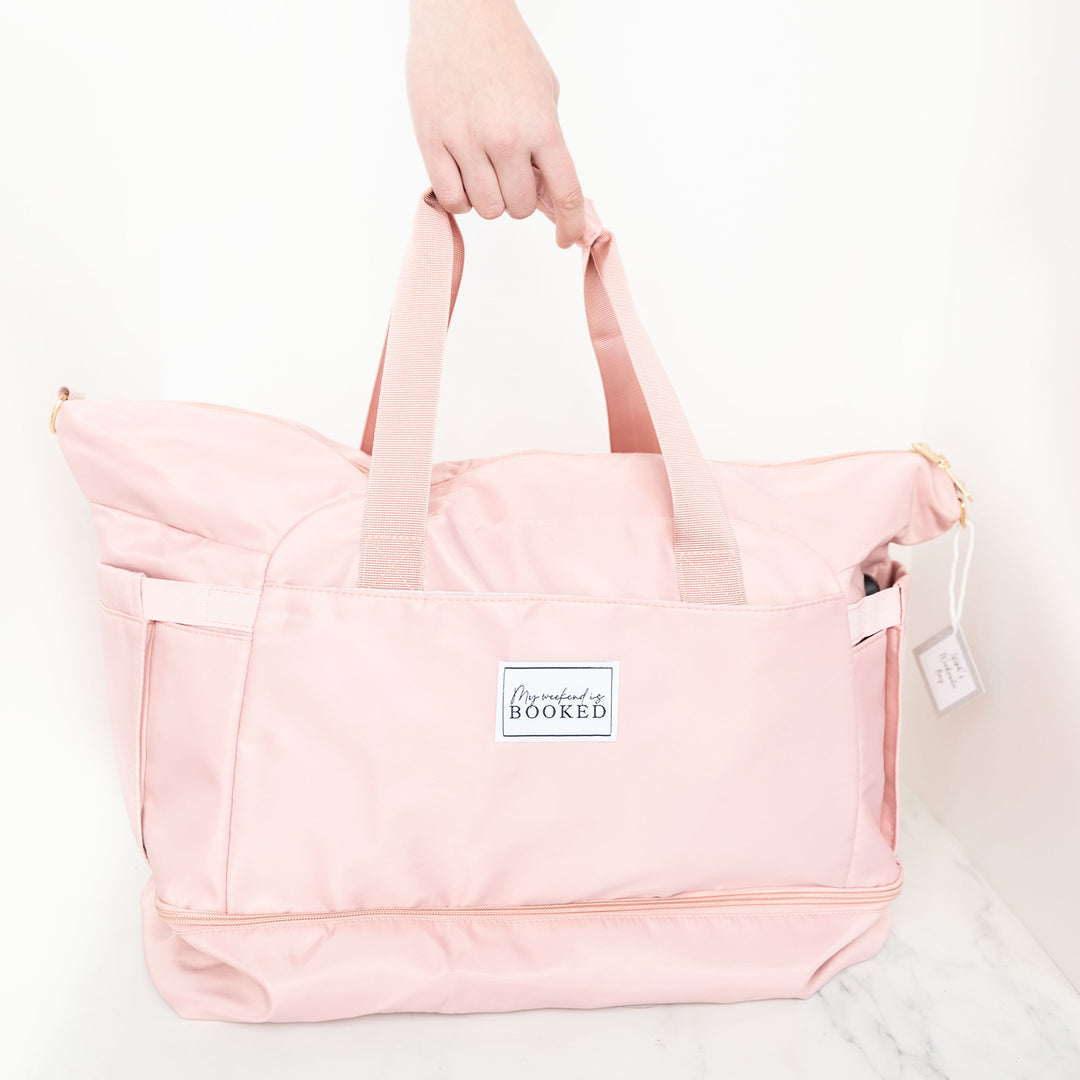 A white hand grips the handle of a pink weekender bag. The bag has a patch on the front that reads "My Weekend is Booked".