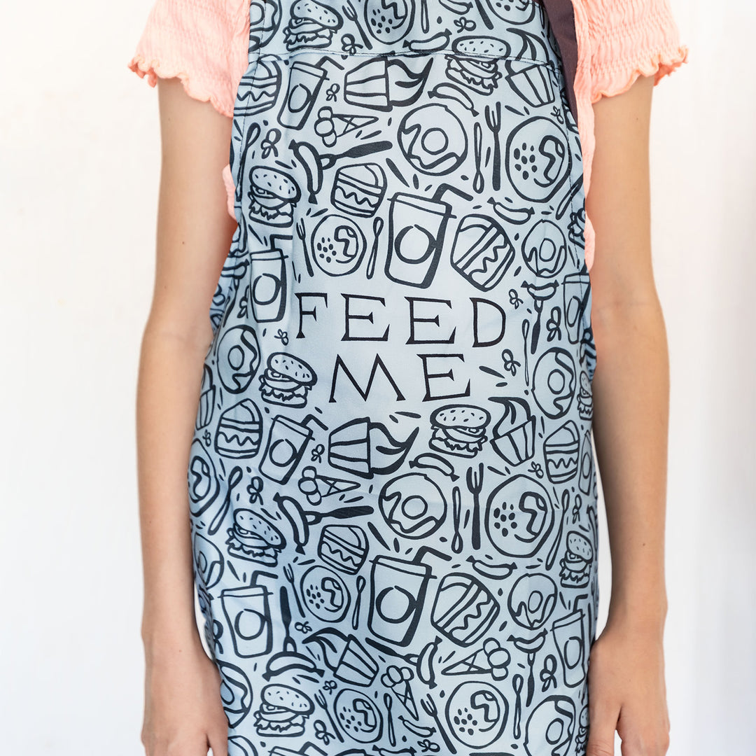 a light blue children's apron with a black pattern across the front of it including sketches of various food and drink. The words FEED ME are in big letters in the middle of the design.