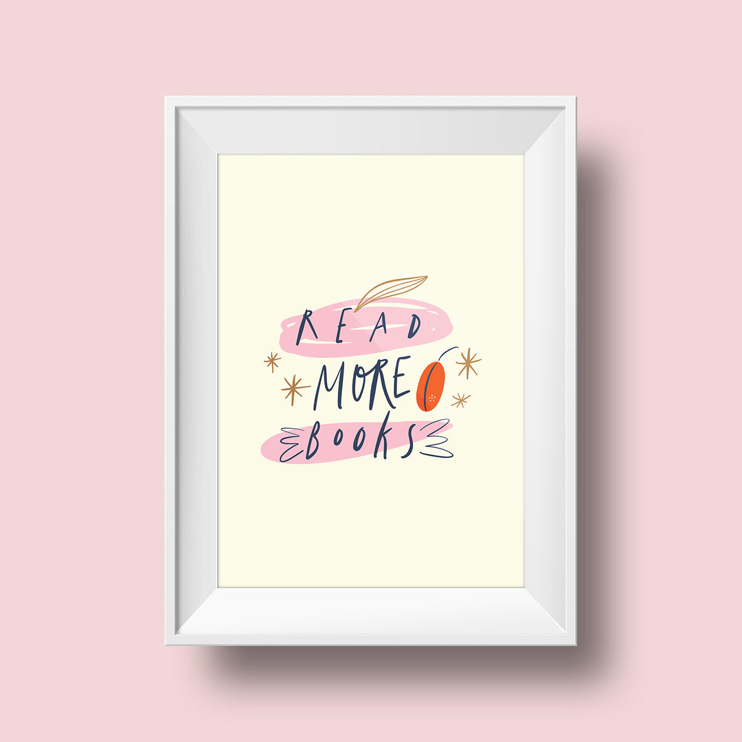 An art print hangs on a pink wall inside a white matted frame. The print features a cream colored background with the words "Read More Books" written in dark blue writing over pink and gold details. Gold stars surround the words.