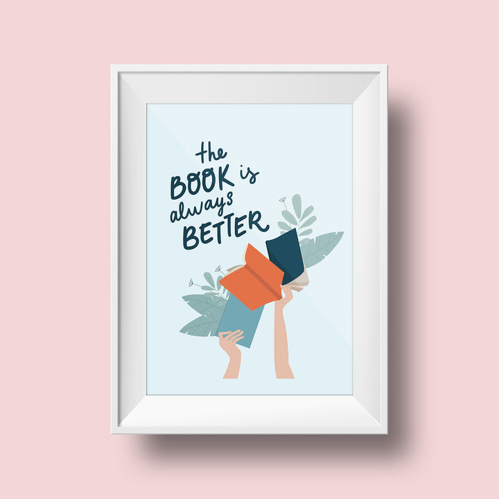 An art print hangs on a pink wall inside a white matted frame. The print features a light blue colored background with the words "The book is always better" written in dark blue script above an illustrated set of hands holding up three multicolored books. Leaves stick out from the pages of the books.