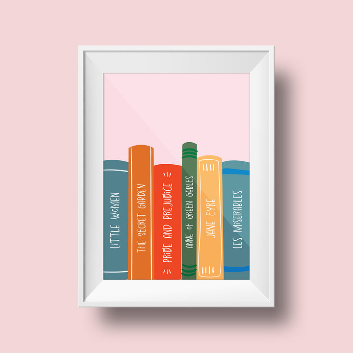 An art print hangs on a pink wall inside a white matted frame. The print features a light pink background with multicolored book spines visible along the bottom of the print. In order, the spines are a dark blue Little Women, an orange The Great Gatsby, a red Pride and Prejudice, a green Anne of Green Gables, a yellow Jane Eyre, and a blue Les Miserables.