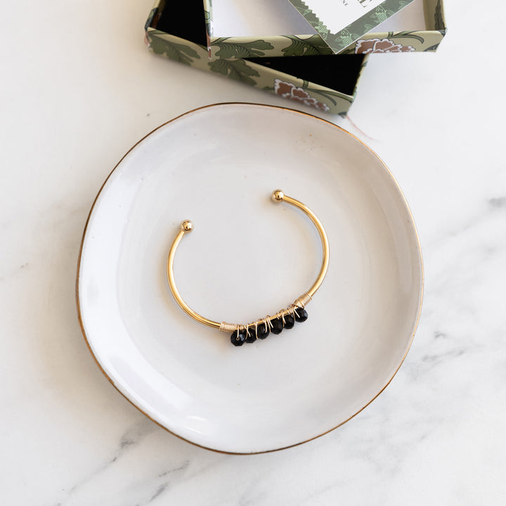 a gold cuff bracelet with black stones wired to the top of it rests on a white a gold plate.