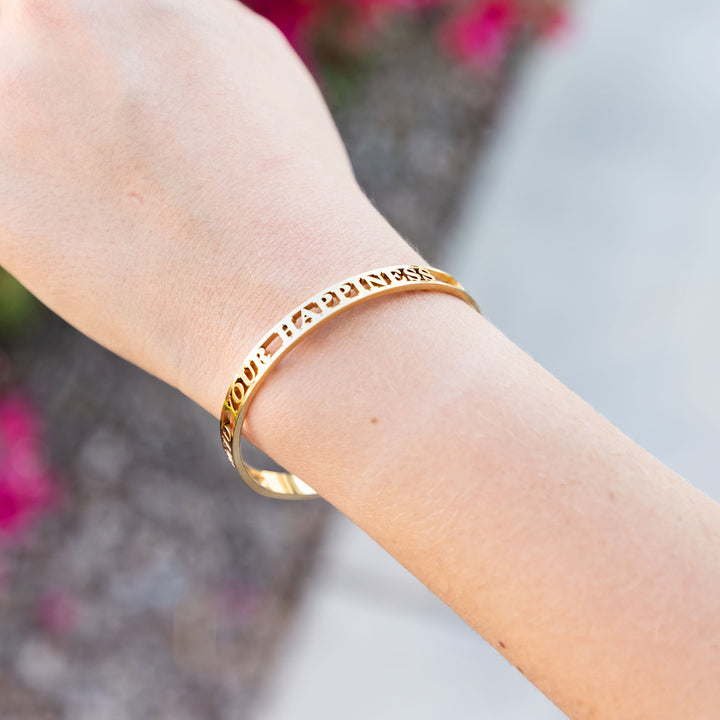 A close-up of a gold bangle bracelet on a person's wrist, engraved with the phrase "FIND YOUR HAPPINESS" against a blurred floral background.