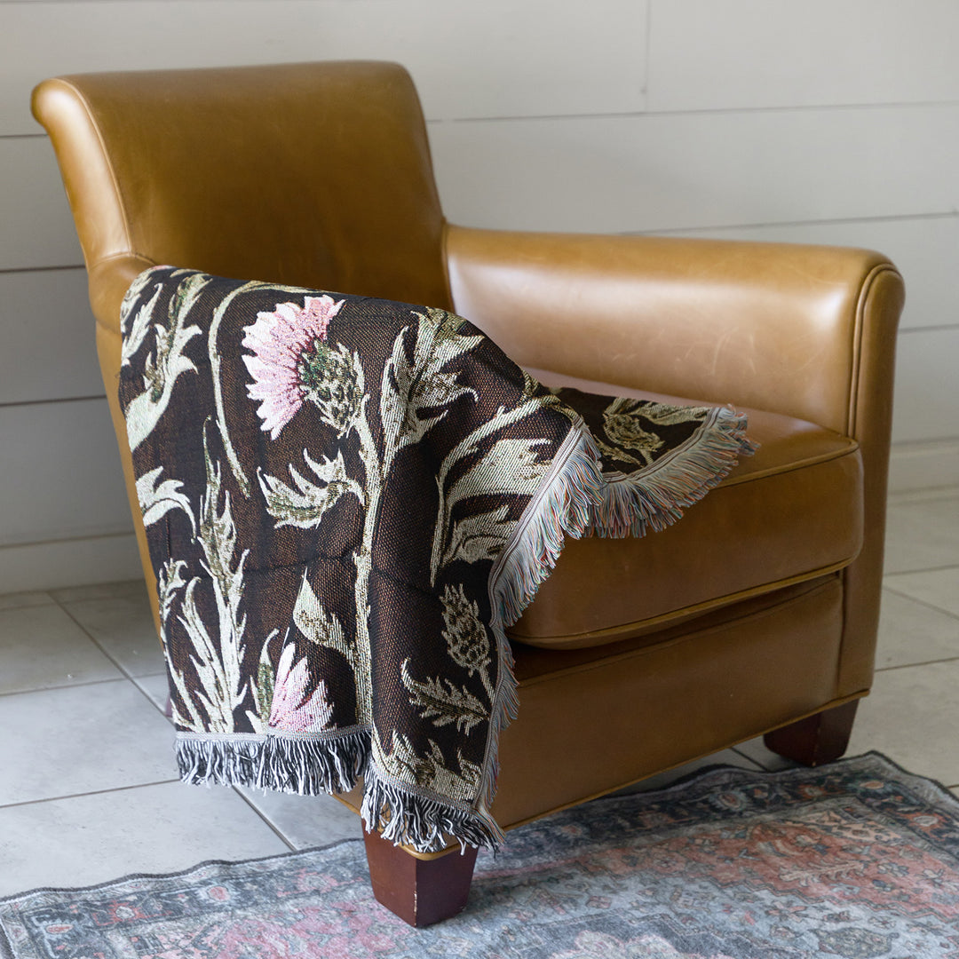 a gorgeous brown woven blanket with tasseled edges stitched with a pink and green thistle flower pattern. Tossed over the arm of a brown leather armchair.