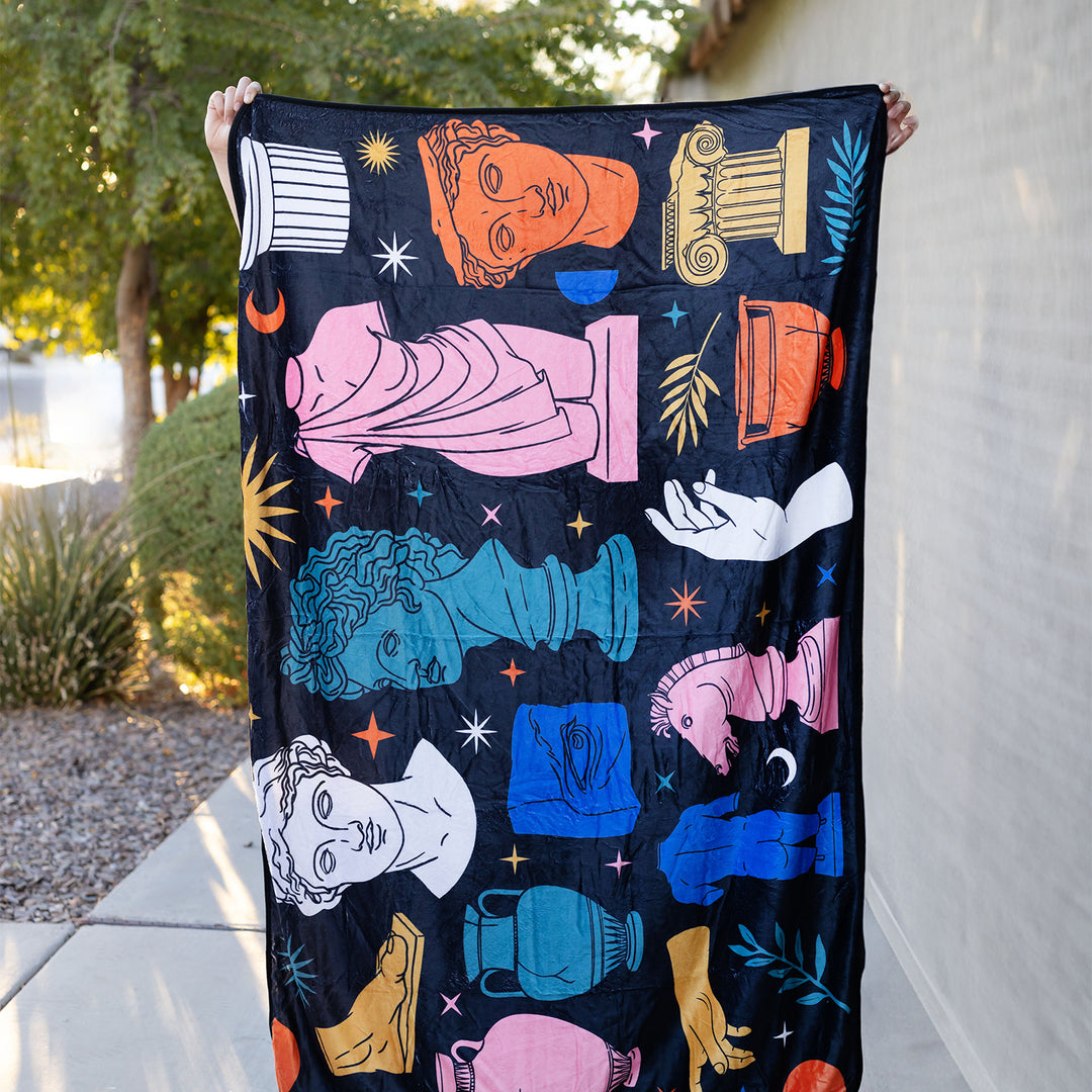 a Greek mythology artwork inspired multi-colored blanket being held up outdoors.