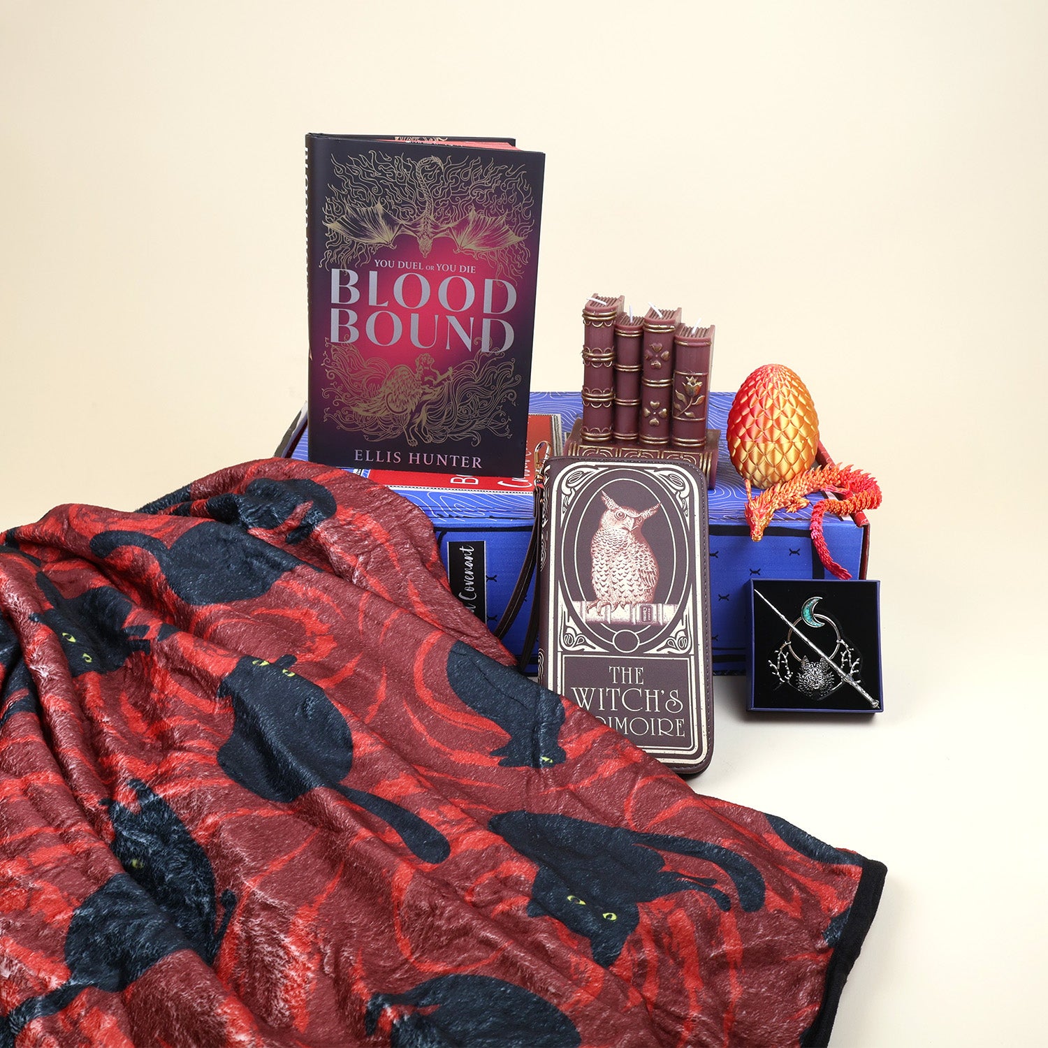 A hardcover special edition of Blood Bound stands on a blue Cursed Covenant OUABC box. Next to the book is a candle shaped like a set of 5 brown books with gold accents, a 3D printed pink/yellow ombre dragon egg and a matching dragon. In front of the box are a red blanket with black cat pattern, a brown leather wallet with an owl that says The Witch's Grimoire, and an open box with a hair pin. The hair pin is a circle with branches, a wolf, and an opal moon around it with a sword piece through the middle.
