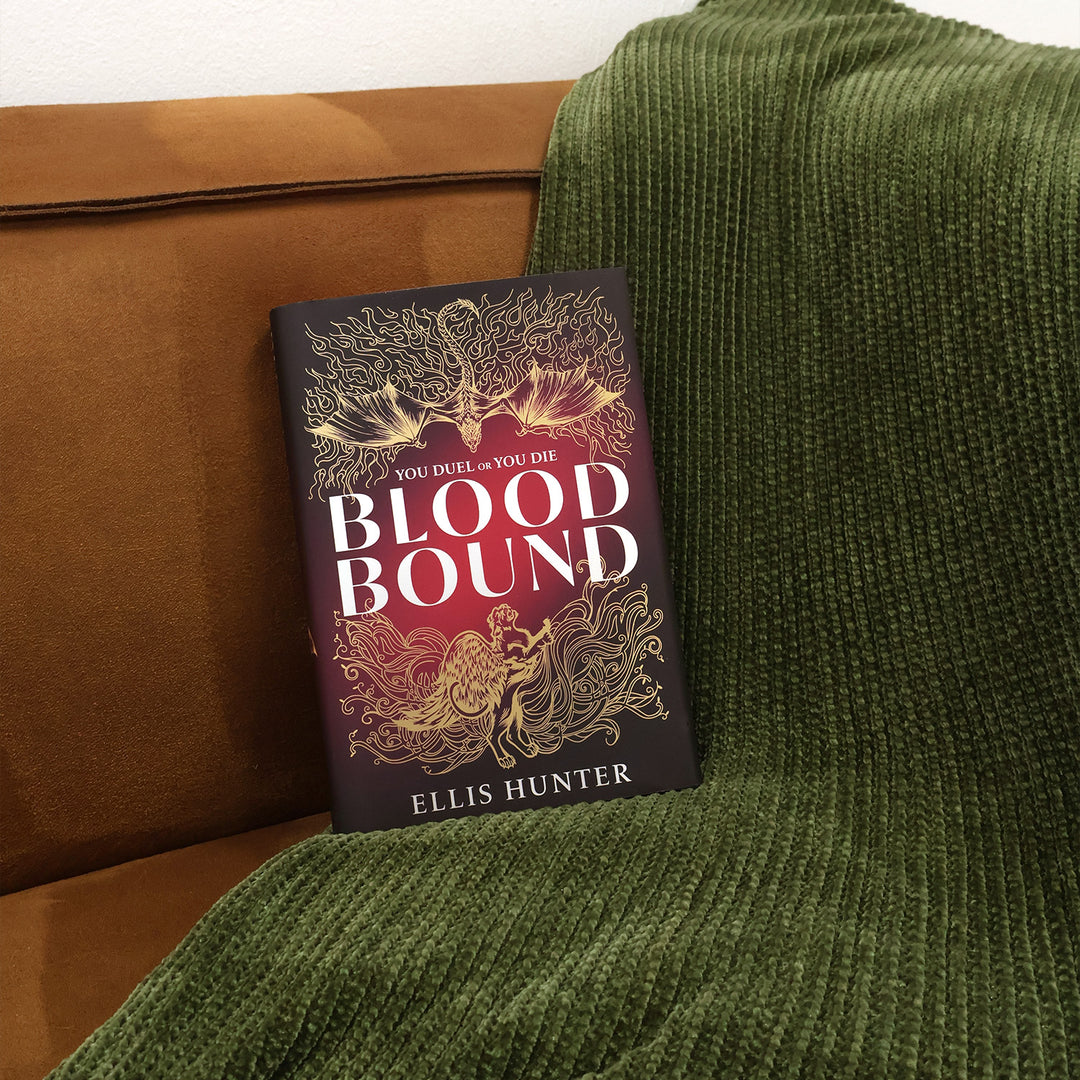 A book titled 'Blood Bound' by Ellis Hunter, featuring a dark red and gold cover, resting on a brown leather couch next to a textured green knit blanket.