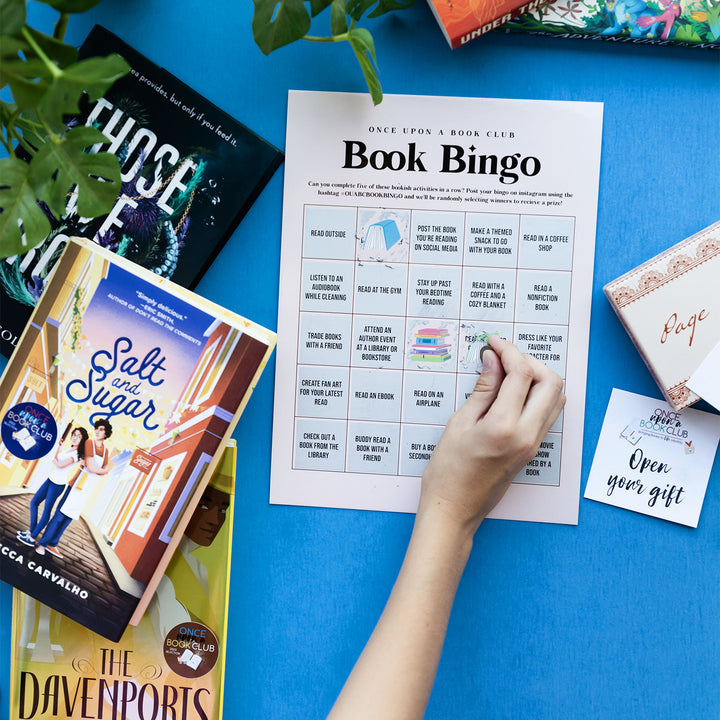 a white hand scratches off a square of Book Bingo next to a stack of books