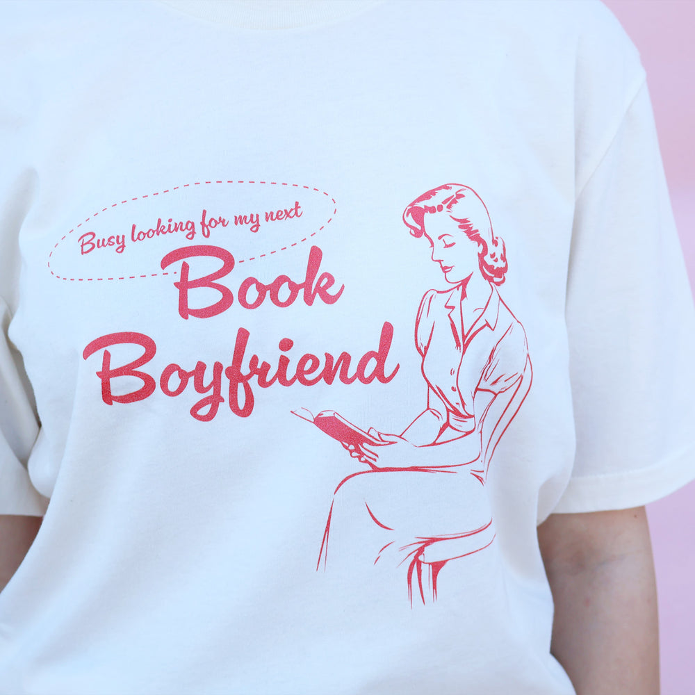 White t-shirt with red text and graphic of a woman reading a book, on a pink background.