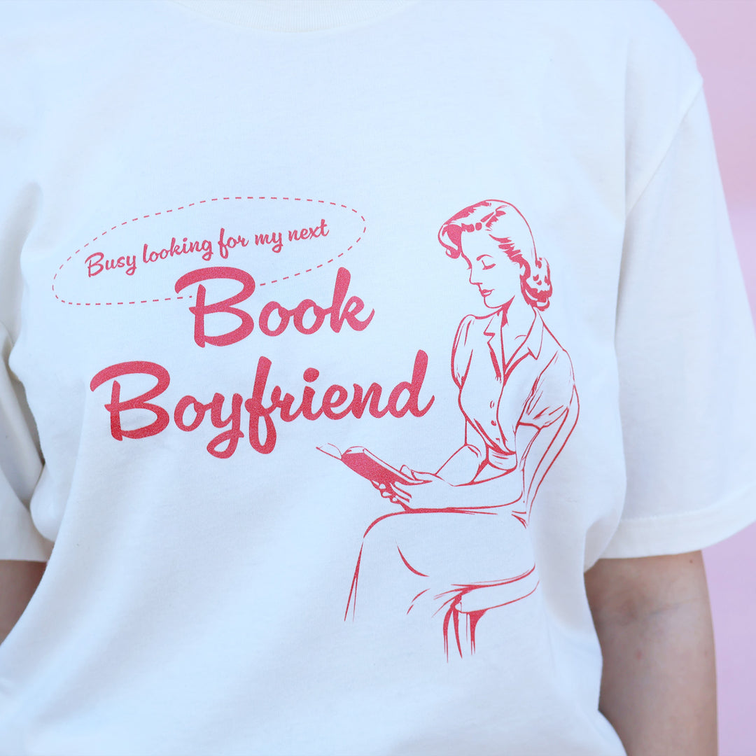 White t-shirt with red text and graphic of a woman reading a book, on a pink background.