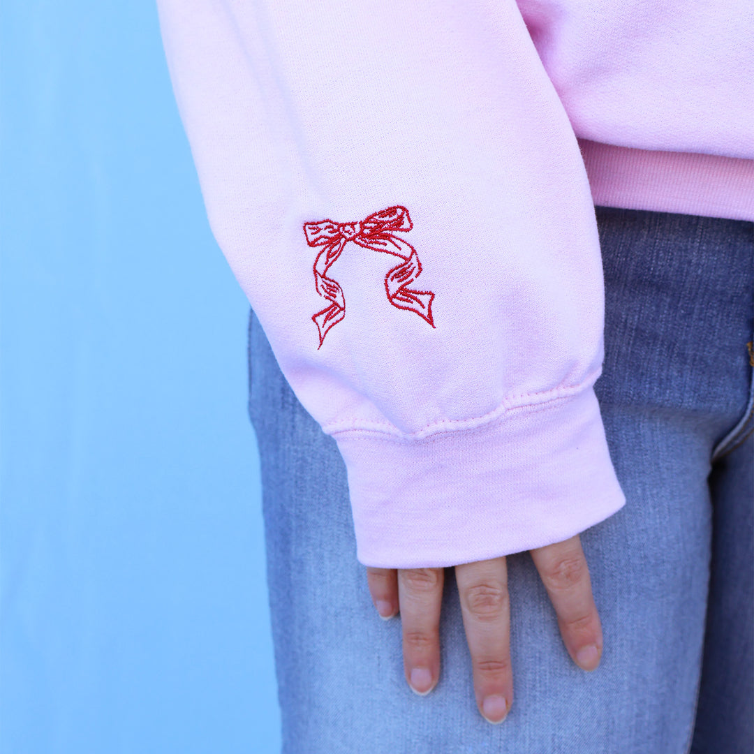 Pink sweatshirt with red embroidered bow design on sleeve against a blue background