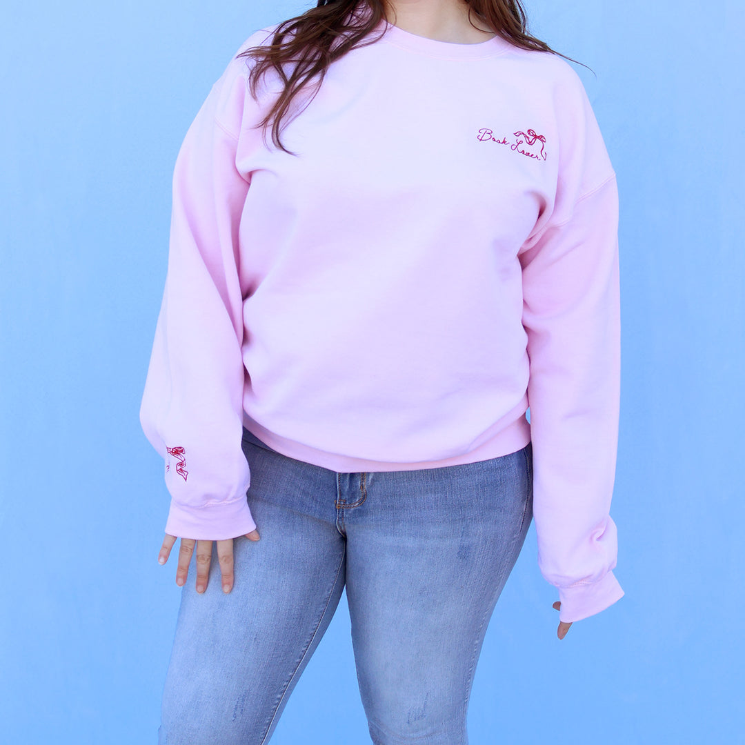 Person wearing a pink sweatshirt with a red embroidered book lover quote and bow against a blue background