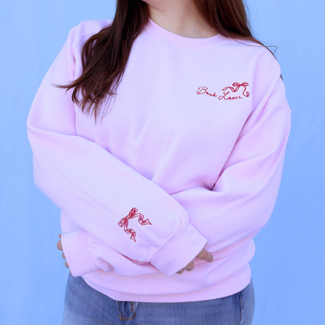 Person wearing a pink sweatshirt with a red embroidered book lover quote and bow against a blue background