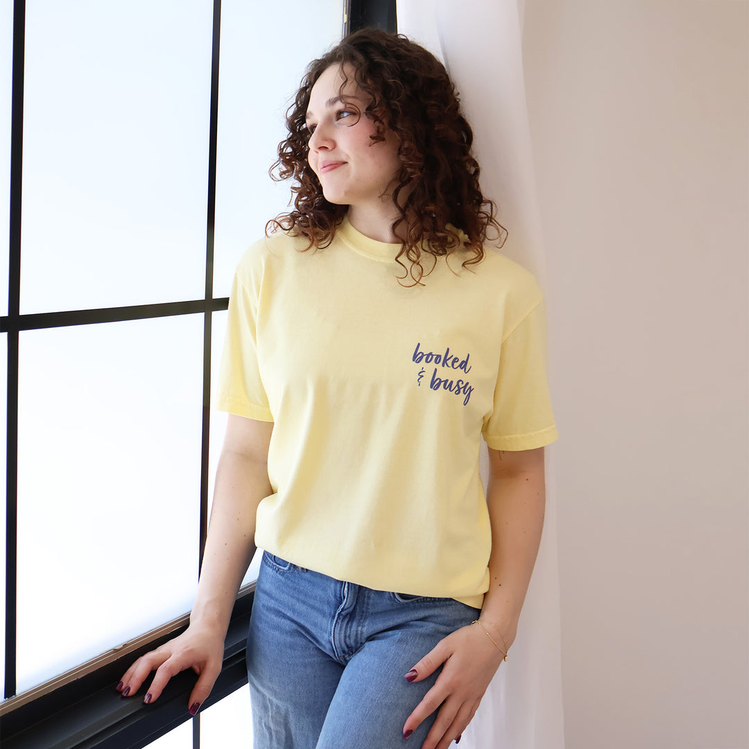 A woman wearing yellow t-shirts with 'Booked & Busy' on the front and 'Ask Me What I'm Reading' on the back surrounded by book-themed doodles.