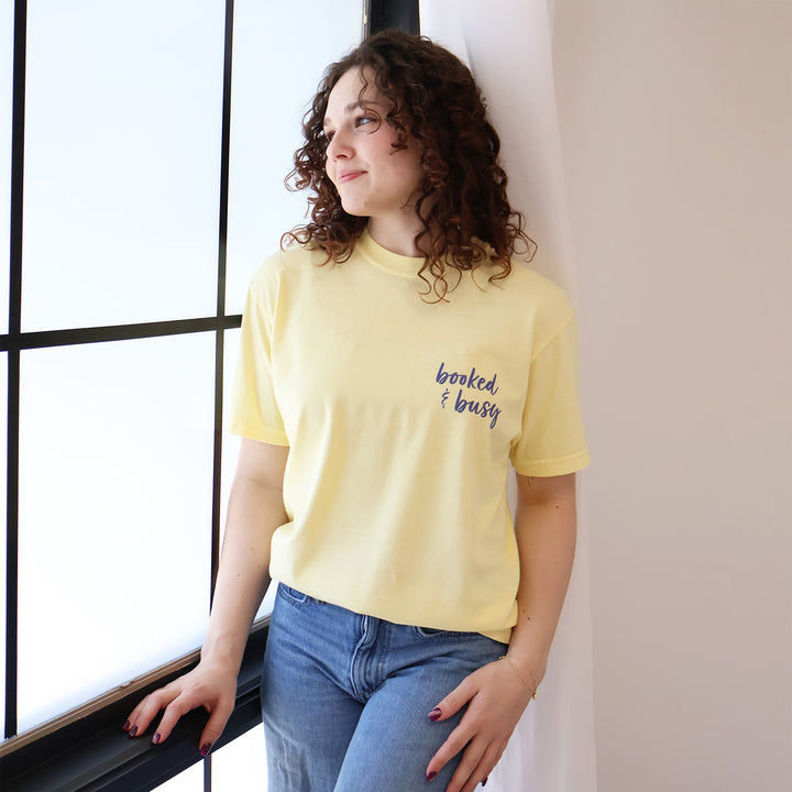 A woman wearing yellow t-shirts with 'Booked & Busy' on the front and 'Ask Me What I'm Reading' on the back surrounded by book-themed doodles.