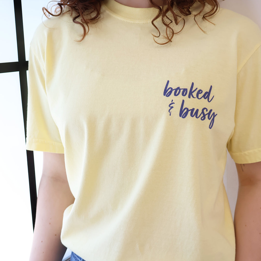 A woman wearing a butter yellow t-shirt with 'Booked & Busy' on the front and 'Ask Me What I'm Reading' on the back surrounded by book-themed doodles.