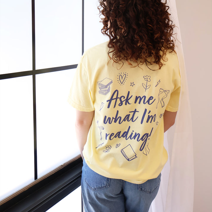 A woman wearing yellow t-shirts with 'Booked & Busy' on the front and 'Ask Me What I'm Reading' on the back surrounded by book-themed doodles.
