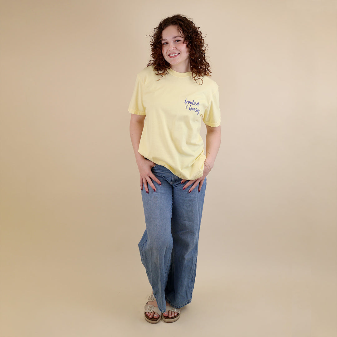 A woman wearing yellow t-shirts with 'Booked & Busy' on the front and 'Ask Me What I'm Reading' on the back surrounded by book-themed doodles.
