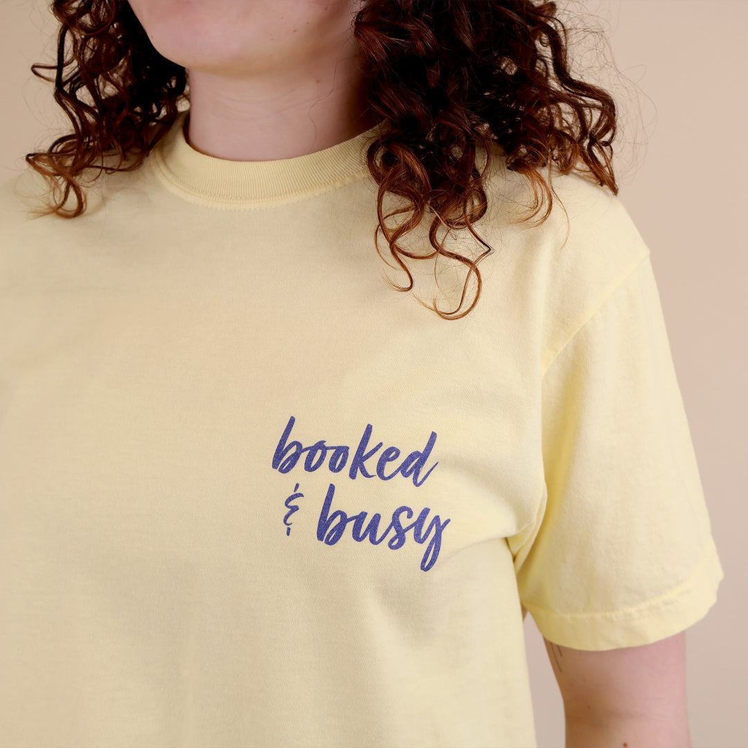 A woman wearing yellow t-shirts with 'Booked & Busy' on the front and 'Ask Me What I'm Reading' on the back surrounded by book-themed doodles.