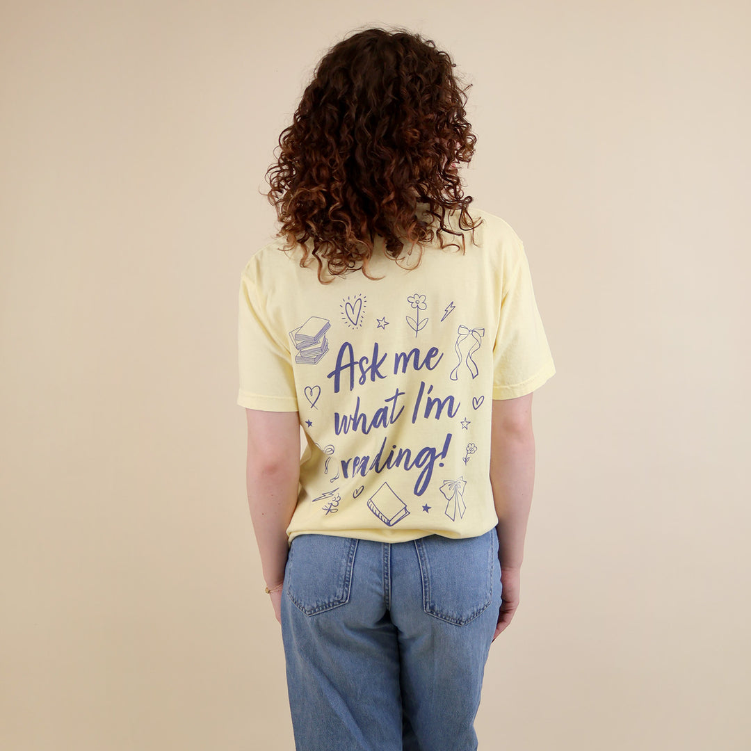 A woman wearing yellow t-shirts with 'Booked & Busy' on the front and 'Ask Me What I'm Reading' on the back surrounded by book-themed doodles.