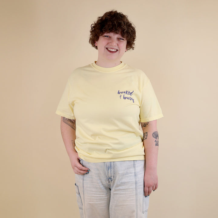 A woman wearing yellow t-shirts with 'Booked & Busy' on the front and 'Ask Me What I'm Reading' on the back surrounded by book-themed doodles.