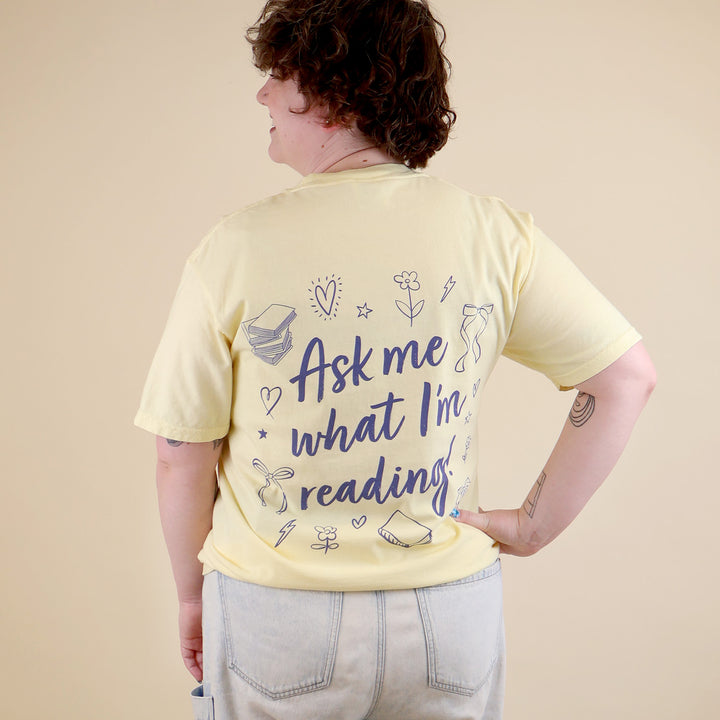 A woman wearing a butter yellow t-shirt with 'Booked & Busy' on the front and 'Ask Me What I'm Reading' on the back surrounded by book-themed doodles.