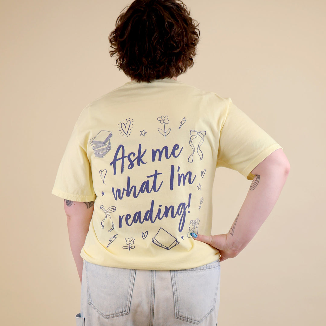 A woman wearing yellow t-shirts with 'Booked & Busy' on the front and 'Ask Me What I'm Reading' on the back surrounded by book-themed doodles.