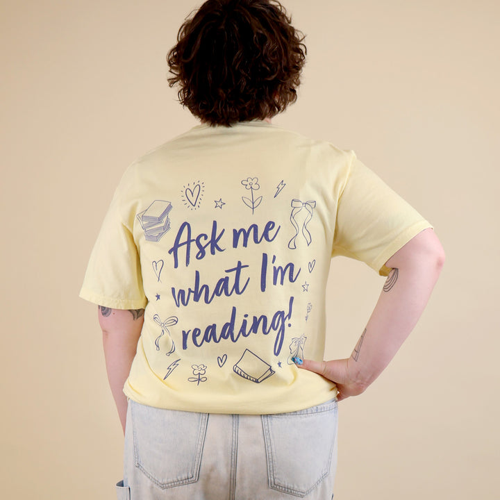 A woman wearing yellow t-shirts with 'Booked & Busy' on the front and 'Ask Me What I'm Reading' on the back surrounded by book-themed doodles.