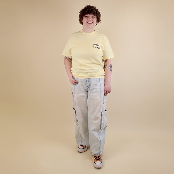 A woman wearing yellow t-shirts with 'Booked & Busy' on the front and 'Ask Me What I'm Reading' on the back surrounded by book-themed doodles.