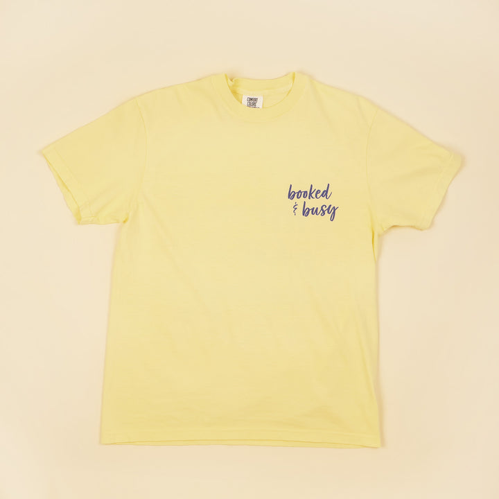 A yellow t-shirt with 'Booked & Busy' on the front and 'Ask Me What I'm Reading' on the back surrounded by book-themed doodles.