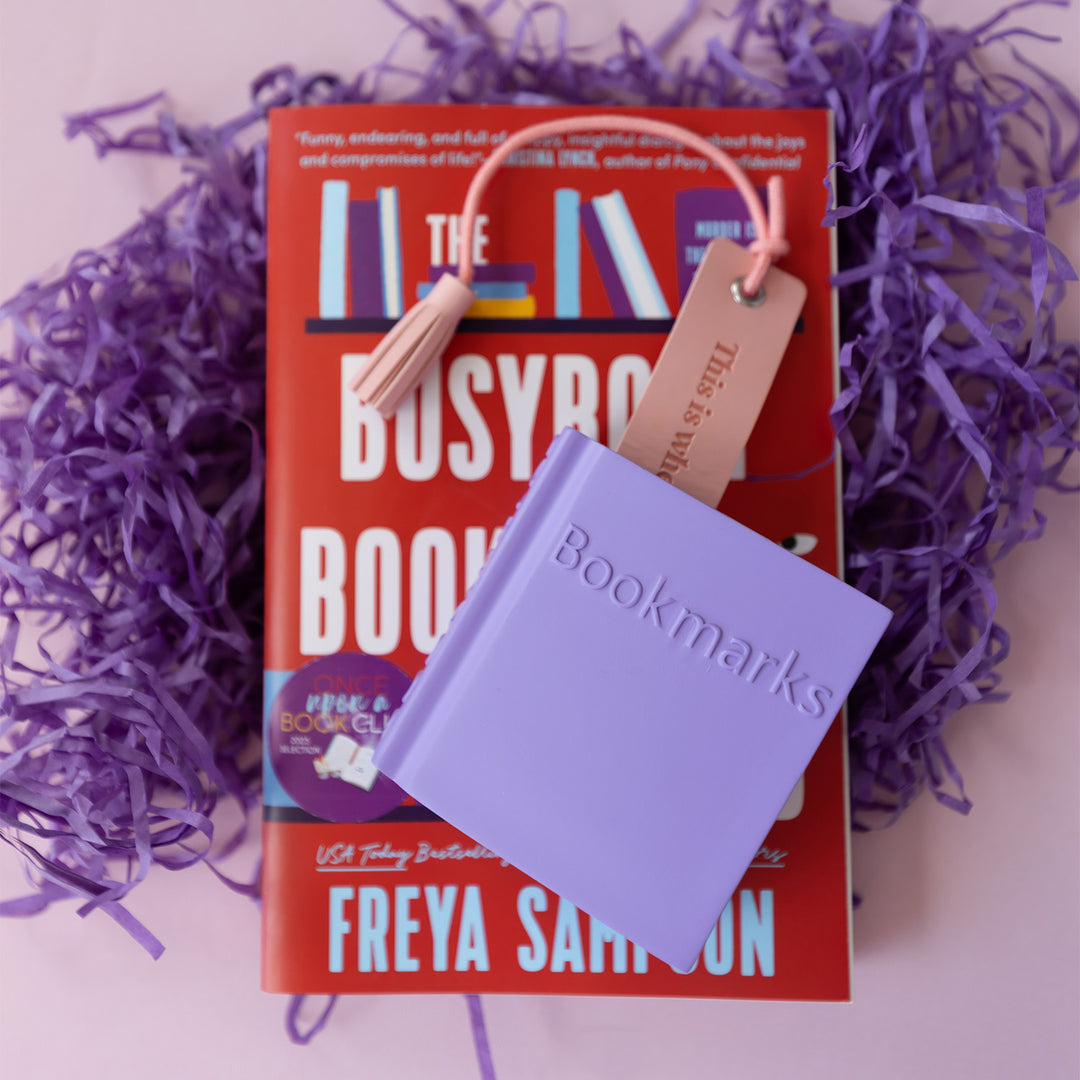 A pink tasseled bookmark sticks out of a short purple container labeled Bookmarks. The container lays on a paperback edition. of The Busybody Book Club surrounded by purple string confetti.