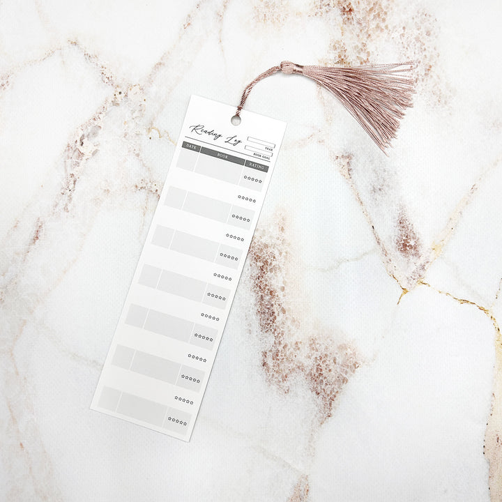 An individual reading log bookmark with a pink tassel. The bookmark has space for the year, book goal, date, book title, and star rating. It sits on a marble background.