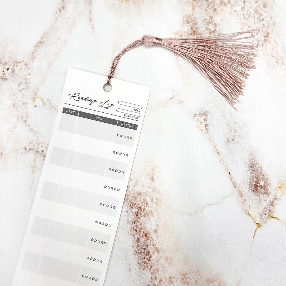 A close up of an individual reading log bookmark with a pink tassel. The bookmark has space for the year, book goal, date, book title, and star rating. It sits on a marble background.