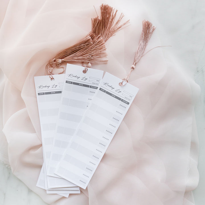 A collection of reading log bookmarks sits on a tulle pink background. The pink tassels of the bookmarks stand up as the bookmark pile splays across the fabric. 