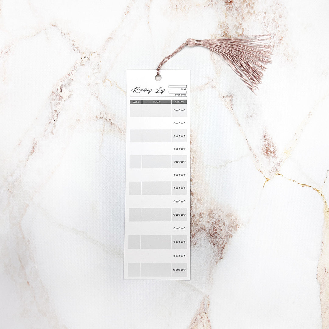 An individual reading log bookmark with a pink tassel. The bookmark has space for the year, book goal, date, book title, and star rating. It sits on a marble background.