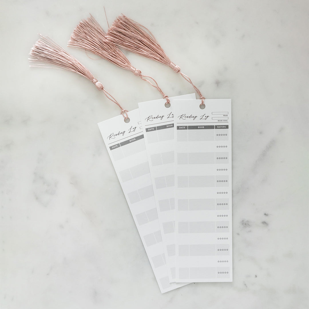 A collection of three Reading Log bookmarks on a white counter. Each bookmark has a pink tassel hanging off the top of it. Bookmarks are equipped with columns for the Date Read, Book Title, and Star Rating. The top front of each bookmark also has a slot for the year the bookmark is tracking and the reading goal for the year.