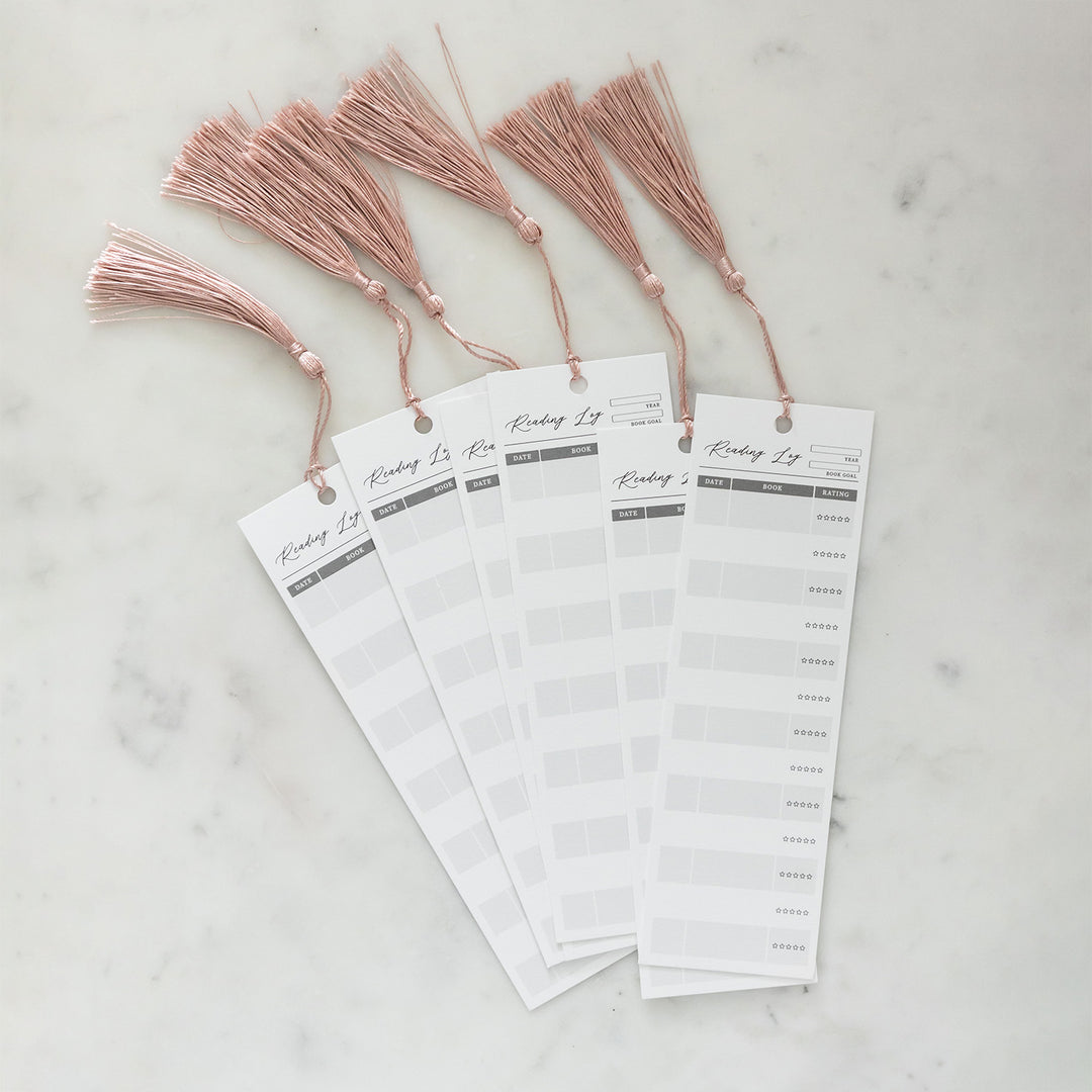 A collection of six Reading Log bookmarks on a white counter. Each bookmark has a pink tassel hanging off the top of it. Bookmarks are equipped with columns for the Date Read, Book Title, and Star Rating. The top front of each bookmark also has a slot for the year the bookmark is tracking and the reading goal for the year.