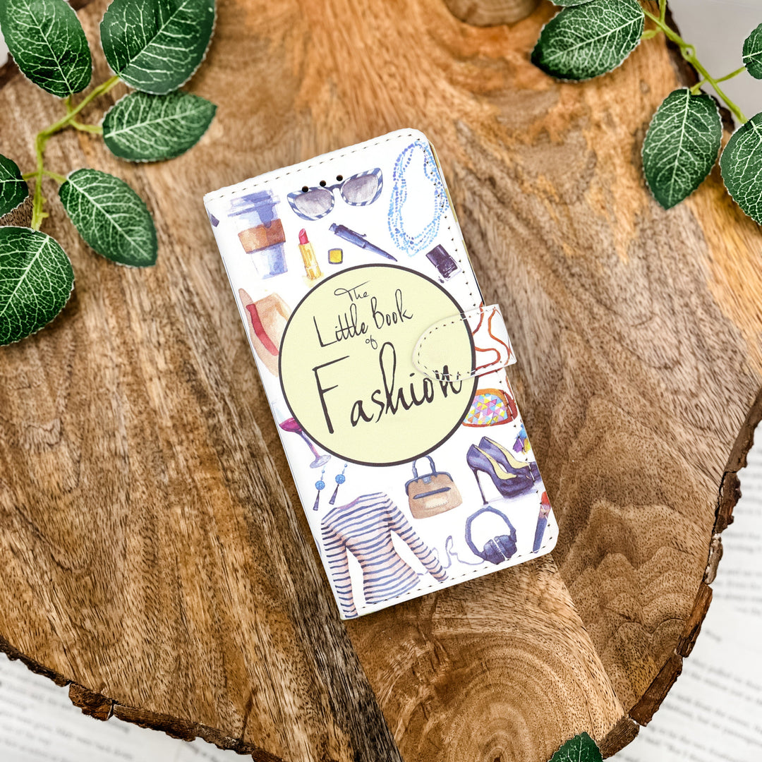Book-shaped phone case titled 'The Little Book of Fashion' on a wooden surface with green leaves.