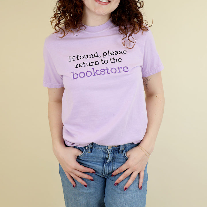 A woman wearing a light purple t-shirt with 'If found, please return to the bookstore' embroidered on it, standing on a beige background.