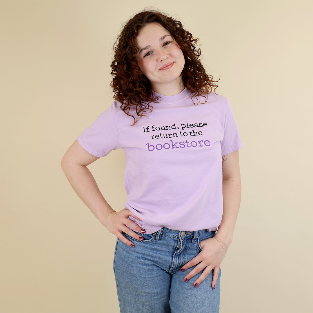 A woman wearing a light purple t-shirt with 'If found, please return to the bookstore' embroidered on it, standing on a beige background.