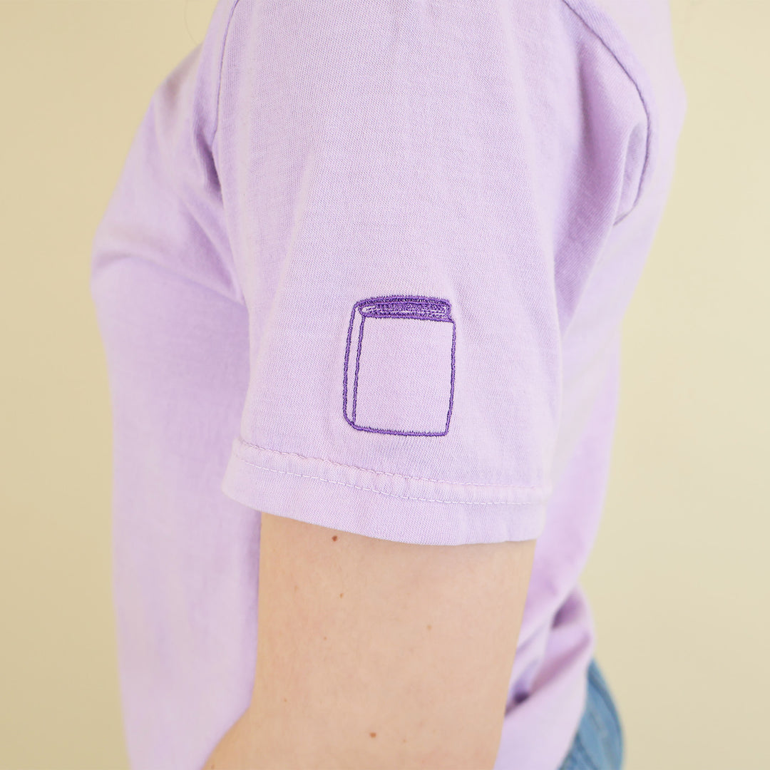 Close-up of a woman wearing a light purple shirt with a book embroidered on the sleeve standing on a beige background